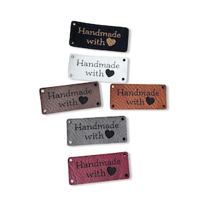 May include: A collection of rectangular tags in various colors, including black, white, brown, and burgundy. Each tag features the words "Handmade with" and a heart symbol. The tags are made of a textured material and have small holes at each corner.