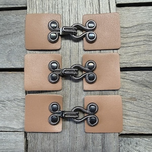 May include: Three sets of tan leather toggle clasps with antique silver-tone metal hardware. Each clasp features a square leather piece and a decorative hook and eye closure. Suitable for coats, jackets, or craft projects.