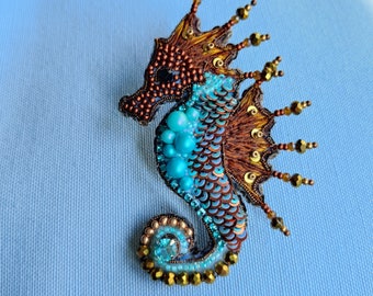 Puede incluir: Un broche de caballito de mar con cuentas, con una corona de hilo dorado y marrón, lentejuelas turquesa y azules y un cristal transparente en la cola.