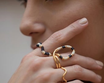 May include: A close-up of a hand wearing two rings. One ring is gold with a wavy design, and the other is a gold ring with a snake-like design, wrapped in black and cream-colored thread. The background is blurred.