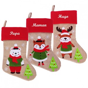 Op de afbeelding: Drie kerstsokken met rode viltmanchetten en jutelichamen. De sokken zijn versierd met geborduurde sneeuwvlokken en hebben een ijsbeer, een vos en een rendier. De sokken zijn gepersonaliseerd met de namen "Papa", "Maman" en "Hugo".