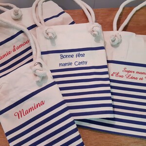 May include: Canvas tote bags with blue and white stripes and rope handles. Several bags are visible, each with embroidered text in red. The text includes phrases like "Mamie d'amour", "Bonne f&ecirc;te mamie Cathy", and "Super mamie".