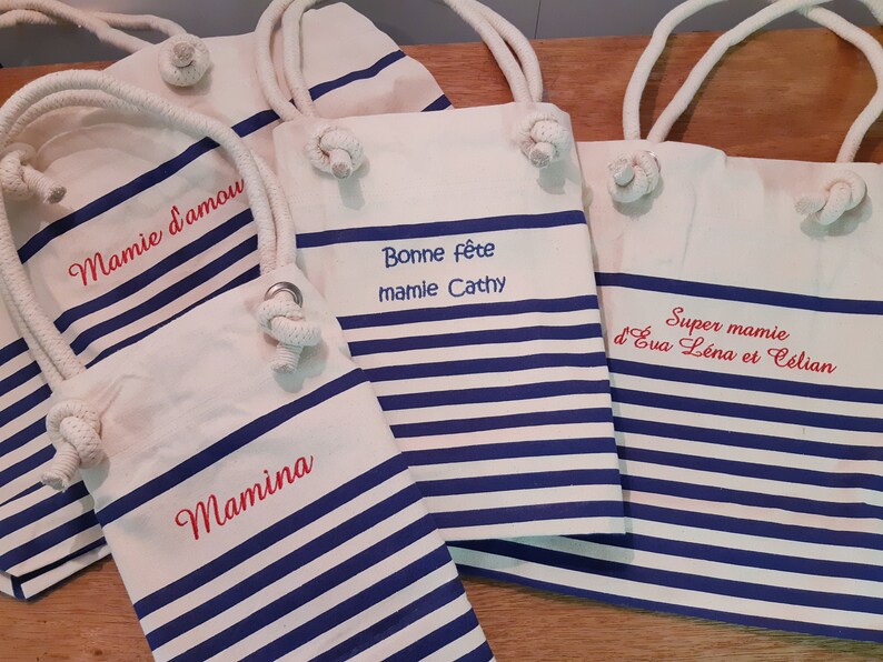 May include: Canvas tote bags with blue and white stripes and rope handles. Several bags are visible, each with embroidered text in red. The text includes phrases like "Mamie d'amour", "Bonne f&ecirc;te mamie Cathy", and "Super mamie".