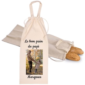 Peut inclure: Un sac à baguette en tissu beige avec une fermeture à cordon et une poignée. Le sac présente le texte imprimé Le bon pain de papi, une photographie d'un adulte et d'un enfant se tenant la main dans un parc, et le nom Margaux en bas.