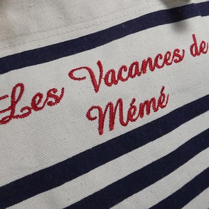 May include: A white tote bag with navy blue stripes and the embroidered text "Les Vacances de M&eacute;m&eacute;" in red. The bag is made of a sturdy fabric, perfect for carrying items.
