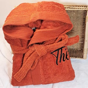 Puede incluir: Bata de baño naranja con capucha y cinturón a juego. La palabra "The" está bordada en negro en la parte delantera. La bata está doblada sobre una superficie blanca, con una bandeja de mimbre en el fondo.