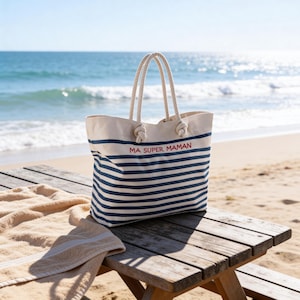 May include: A canvas tote bag with navy blue horizontal stripes and rope handles. The bag has the text "MA SUPER MAMAN" in red. A beige beach towel rests on a wooden picnic table, with the ocean in the background.