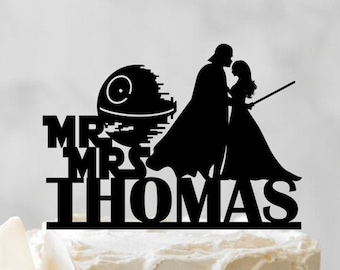 May include: A white wedding cake with a black silhouette of Darth Vader and Princess Leia kissing on top of the cake. The cake topper also includes the Death Star and the text "Mr. & Mrs. Thomas".