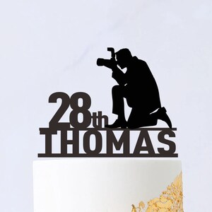 Puede incluir: Silueta negra de una persona sosteniendo una cámara con el texto "28th THOMAS" en un pastel blanco con detalles dorados.