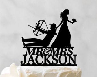 May include: A black silhouette cake topper of a bride and groom. The groom is holding a bow and arrow and the bride is holding a bouquet. The topper says "Mr. & Mrs. Jackson" in black lettering.