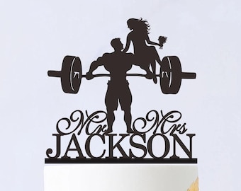 May include: Black silhouette of a couple on a wedding cake topper. The groom is lifting a barbell with the bride sitting on it. The text "Mr. & Mrs. JACKSON" is below the couple.