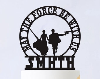 May include: A black cake topper featuring a silhouette of a couple with weapons, encircled by the phrase "MAY THE FORCE BE WITH US" and the name "SMITH". The topper is placed on a white cake with gold leaf accents and a white flower.