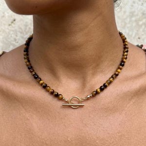 May include: A beaded necklace featuring round, polished tiger's eye stones in shades of brown and gold. The necklace is secured at the front with a gold-toned metal toggle clasp, consisting of a circular ring and a horizontal bar closure mechanism.