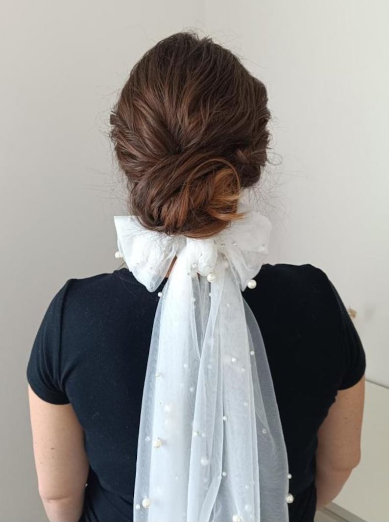 Peut inclure: Un accessoire de cheveux en tulle blanc avec des fausses perles, noué en nœud. L'accessoire est attaché à un chignon de cheveux bruns. La personne porte un haut noir. Le tulle retombe dans le dos.
