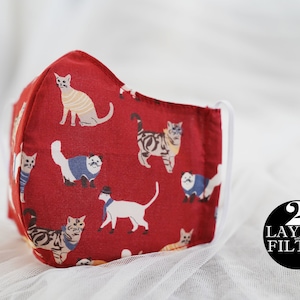 Op de afbeelding: Rood stoffen gezichtsmasker met een herhalend patroon van katten in verschillende outfits. Het masker heeft witte elastische oorlussen en een witte binnenkant. De tekst "2 Layers Filter" staat in een zwarte cirkel.