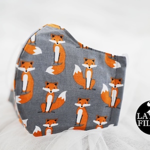 Op de afbeelding: Een grijs stoffen mondkapje met een herhalend patroon van oranje vossen. Het masker heeft twee lagen en een filter. De tekst "2 LAYERS FILTER" is gedrukt op een zwarte cirkel in de rechterbenedenhoek van de afbeelding.