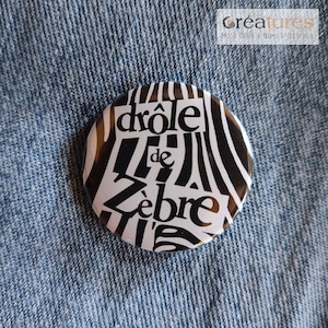 Può includere: Un bottone bianco e nero con il testo "drôle de zèbre" in un carattere stilizzato. Il bottone ha un motivo a righe di zebra.