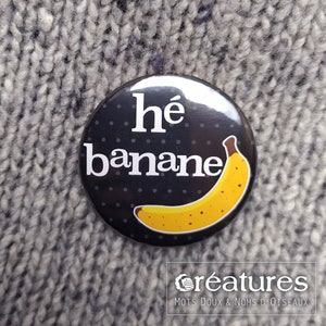 May include: A black button with white polka dots and the text "hé banane" in white. A yellow banana with brown spots is pictured on the button.