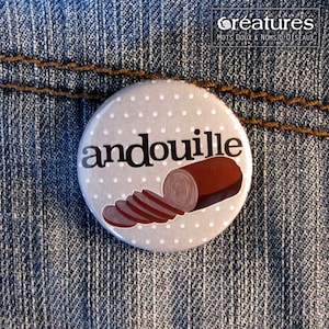 May include: A round button with the word "andouille" in black, over a light gray background with white polka dots. A brown illustration of sliced sausage is on the right. The button is on a blue denim background.