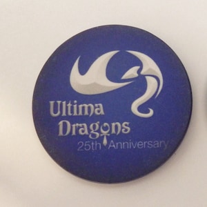 May include: Two blue round tokens with white designs. One token features a white dragon with the text "Ultima Dragons 25th Anniversary". The other token features a white ankh symbol with the text "25 years of Ultima Dragons 1992 2017".
