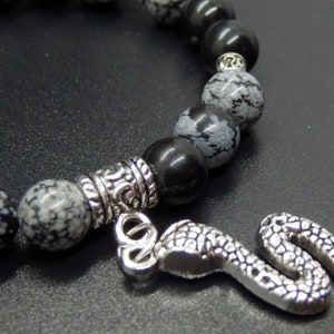 Könnte beinhalten: Ein Perlenarmband mit schwarzen und Schneeflocken-Obsidian-Perlen, silberfarbenen Abstandshaltern und einem Schlangenanhänger. Das Armband ist für das Handgelenk konzipiert.