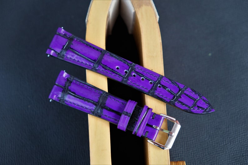 Puede incluir: Dos correas de reloj moradas con estampado de cocodrilo. Una correa tiene una hebilla negra y la otra una hebilla plateada.