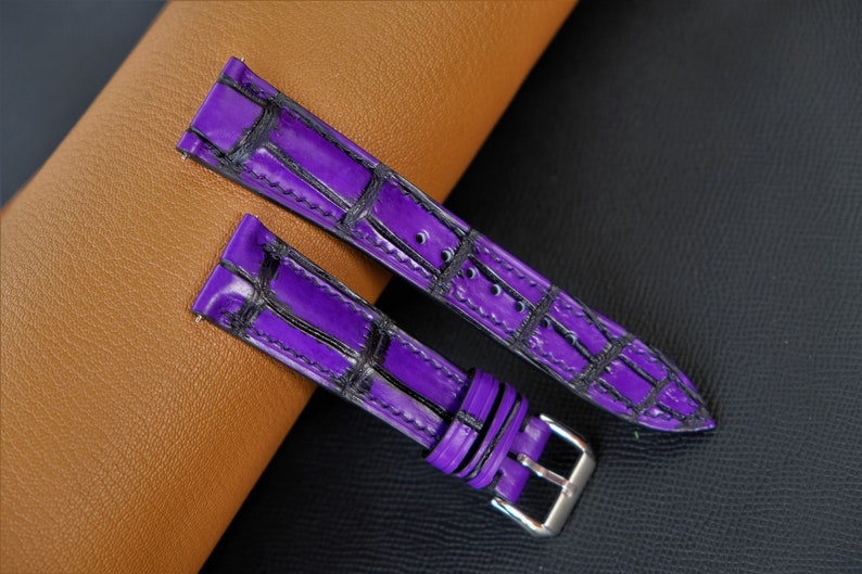 Puede incluir: Dos correas de reloj de cuero morado con un patr&oacute;n de cocodrilo. Las correas tienen costuras negras y se muestran sobre una superficie de cuero marr&oacute;n.