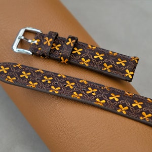 May include: A brown watch strap with a repeating gold floral pattern. The strap has a silver buckle and is displayed on a brown leather surface. The watch strap is a stylish accessory.