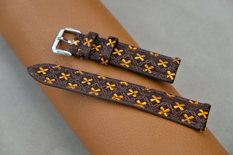 May include: A brown watch strap with a repeating gold floral pattern. The strap has a silver buckle and is displayed on a brown leather surface. The watch strap is a stylish accessory.