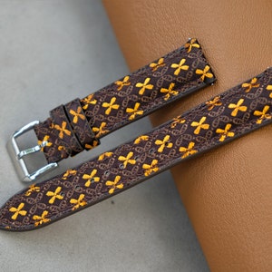 May include: Two-piece watch strap with a brown base and a repeating pattern of yellow cross-like shapes. The strap has a silver buckle and is designed for a watch. The background is a neutral gray and brown.