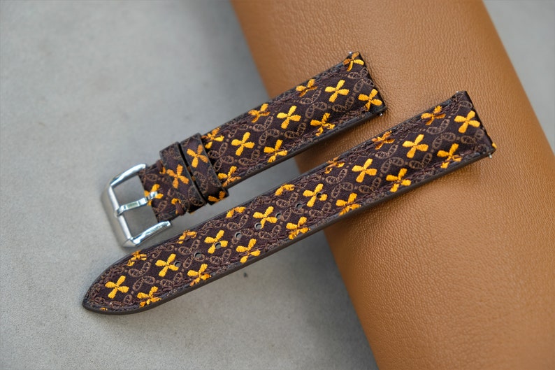 May include: Two-piece watch strap with a brown base and a repeating pattern of yellow cross-like shapes. The strap has a silver buckle and is designed for a watch. The background is a neutral gray and brown.