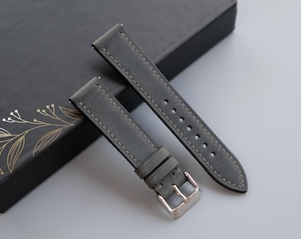 May include: Grey leather watch straps with silver-tone buckles. The straps are stitched with a contrasting thread and are displayed on a dark grey box with gold floral accents. The watch bands are new.