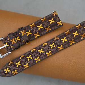 May include: Brown watch strap with a repeating pattern of gold-colored crosses and chain links. The strap has a silver-colored buckle and is displayed on a brown leather surface. The watch band is a stylish accessory.