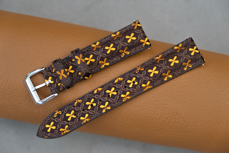 May include: Brown watch strap with a repeating pattern of gold-colored crosses and chain links. The strap has a silver-colored buckle and is displayed on a brown leather surface. The watch band is a stylish accessory.
