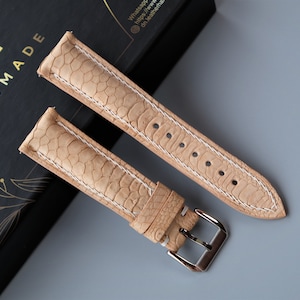 Könnte beinhalten: Zwei hellbraune Lederarmbänder mit strukturiertem Muster und weißen Nähten. Eines der Armbänder hat eine silberne Schnalle. Die Armbänder werden auf einer schwarzen Schachtel mit goldfarbenem Schriftzug "HANDMADE" präsentiert.