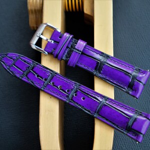 Puede incluir: Dos correas de reloj moradas con un patr&oacute;n de cocodrilo negro. Las correas son de cuero y tienen una hebilla plateada.