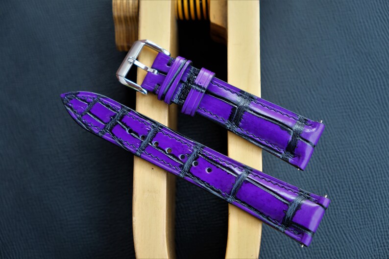Puede incluir: Dos correas de reloj moradas con un patr&oacute;n de cocodrilo negro. Las correas son de cuero y tienen una hebilla plateada.