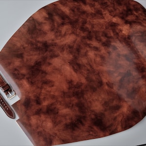 Peut inclure: Une grande pièce de cuir marron de forme irrégulière avec une surface texturée, ainsi que deux bracelets de montre en cuir marron avec des boucles argentées. Le cuir a un aspect marbré, avec des tons bruns plus foncés et plus clairs.