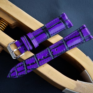 Puede incluir: Dos correas de reloj de cuero morado con un patr&oacute;n de cocodrilo negro. Las correas est&aacute;n en una herramienta de correa de reloj de madera.