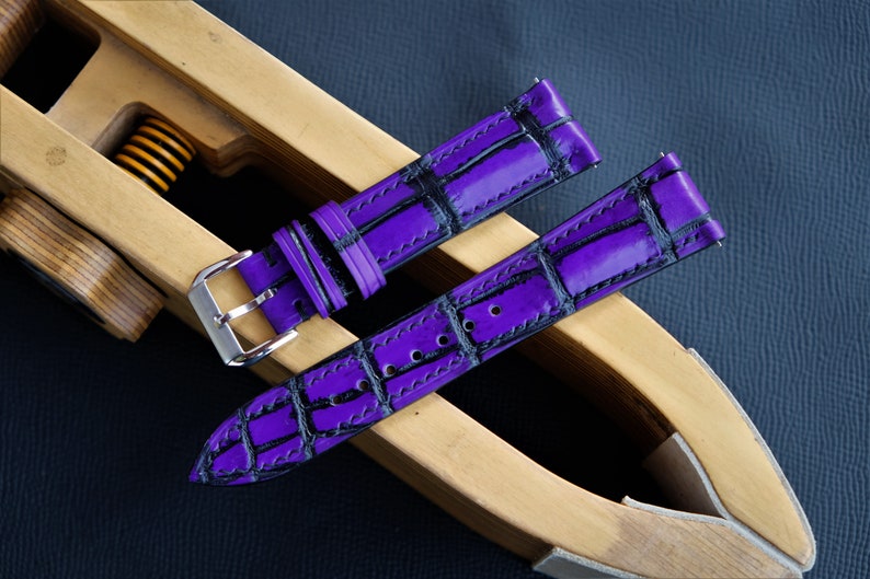 Puede incluir: Dos correas de reloj de cuero morado con un patr&oacute;n de cocodrilo negro. Las correas est&aacute;n en una herramienta de correa de reloj de madera.