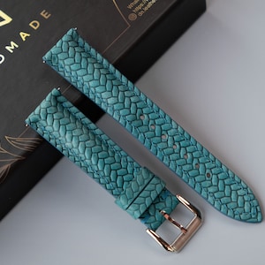Puede incluir: Dos correas de reloj de cuero azul turquesa con un patrón tejido. Las correas están dispuestas sobre una caja negra con letras doradas que dicen "HANDMADE".