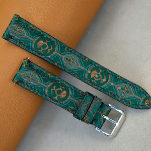 May include: Two-piece watch strap in teal green with a repeating gold pattern. The strap has a silver buckle and is made of a textured fabric. The design features geometric and floral elements.