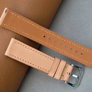 May include: Two leather watch straps in different shades of brown. The straps have contrasting stitching and are laid out on a brown surface.