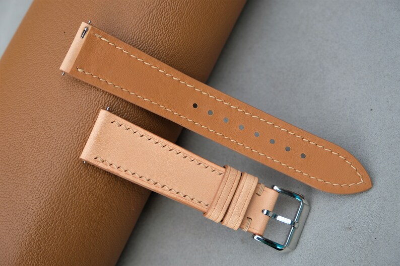 May include: Two leather watch straps in different shades of brown. The straps have contrasting stitching and are laid out on a brown surface.
