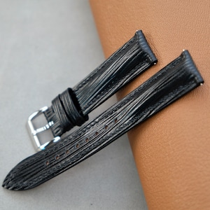 May include: Two black leather watch straps with a textured pattern. The straps are laid out on a brown surface.