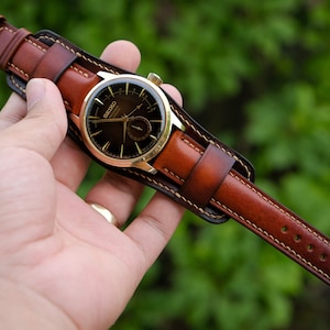 May include: A brown leather watch strap with a gold watch face. The strap is stitched with a light brown thread and has a pointed end.