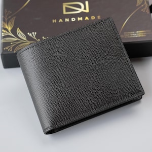 May include: A dark grey leather wallet with a textured finish. The wallet is rectangular and folded, with a box in the background that reads "HANDMADE". The box is dark brown with gold leaf detailing.