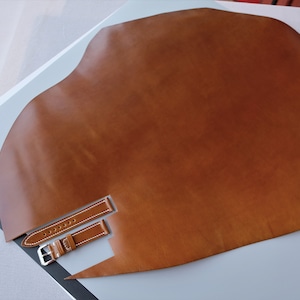 May include: A large piece of brown leather with a watch strap and buckle. The watch strap is brown with white stitching and a silver buckle. The leather is a warm brown color and appears to be smooth.