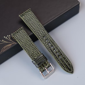 Könnte beinhalten: Olivgrüne Uhrenarmbänder mit einem strukturierten, reptilienartigen Muster. Die Armbänder werden auf einer schwarzen Schachtel mit goldenen floralen Akzenten präsentiert. Die Schnalle ist silberfarben.