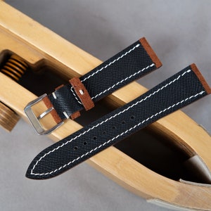 Puede incluir: Dos correas de reloj de cuero negro con costuras blancas. La correa de arriba tiene un forro de ante marr&oacute;n. Las correas est&aacute;n en una herramienta de correa de reloj de madera.